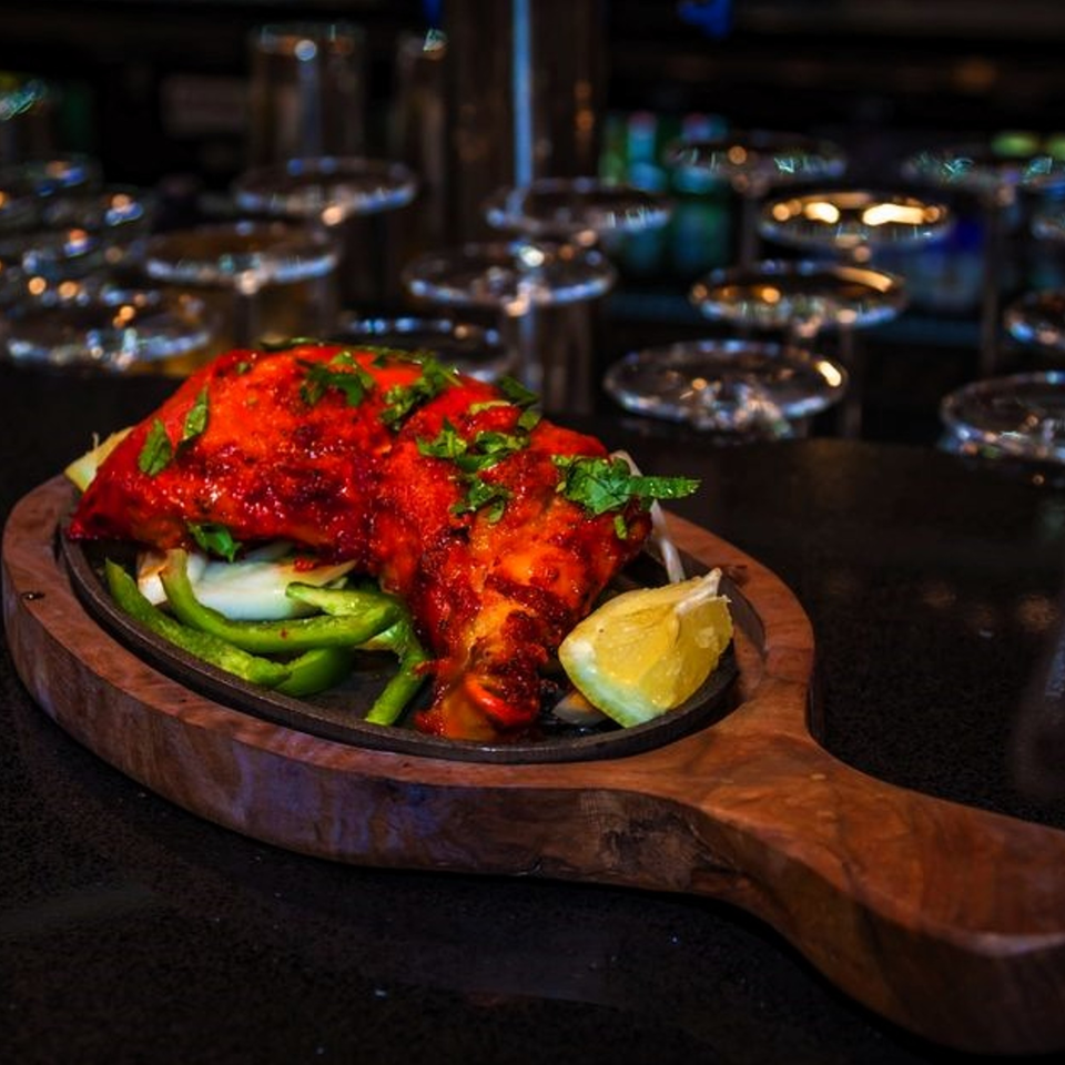 Indian tandoori chicken, garnished with cilantro, onions, bell peppers, and a lemon wedge on a wooden platter.