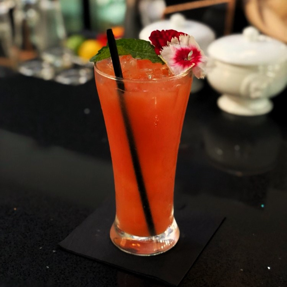 A vibrant orange cocktail with mint and two pink flowers, served in a tall glass on a black coaster.