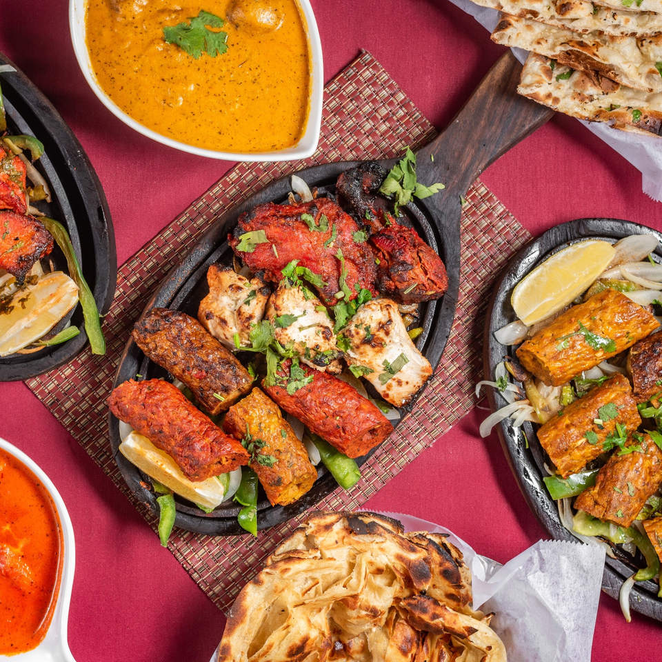 Overhead view of various Indian dishes including tandoori chicken, kebabs, naan, and curry on a red tablecloth.